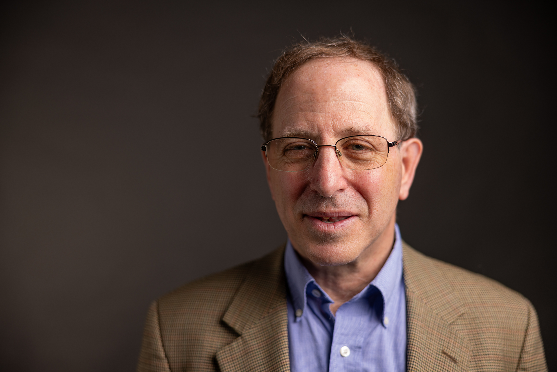 White middle-aged man wearing glasses and blue collared shirt with a beige sport coat