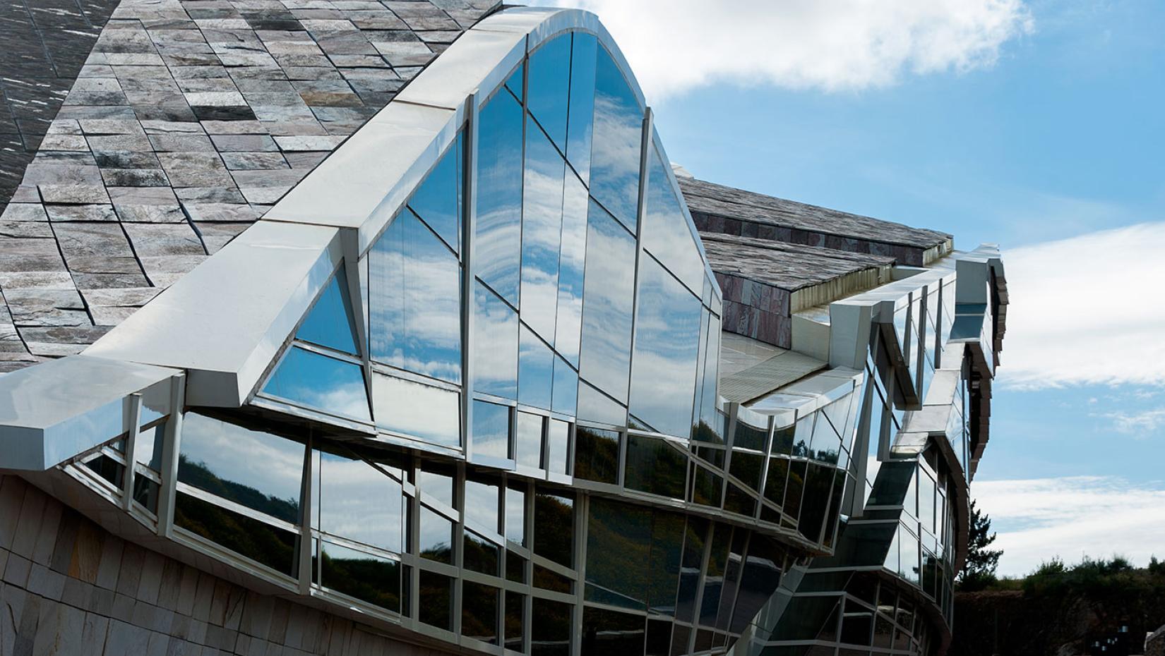 Detail view of curving building with wall of windows reflecting a bright blue sky and clouds.