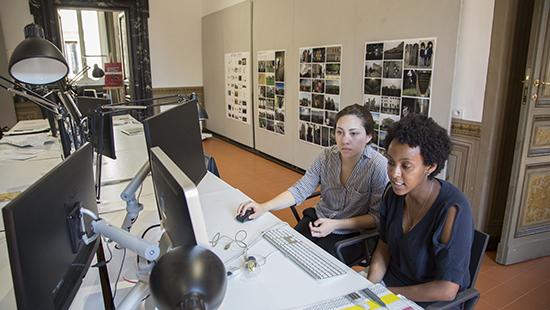 two students in the foreground looking at a computer screen with pinned projects on the background wall