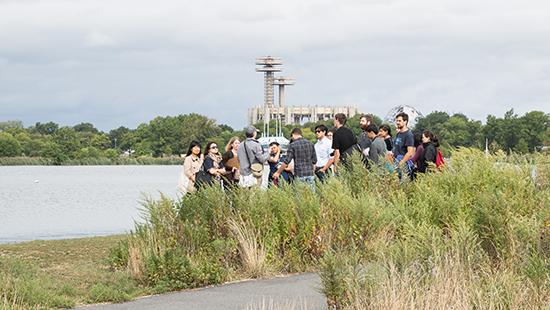 a group of people standing in the grass near water with the World's Fair grounds in the distance