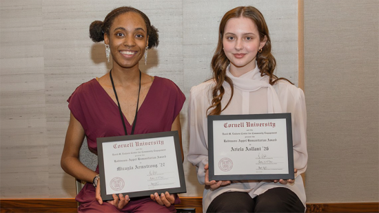 Two individulas seated next to each other holding award plaques.