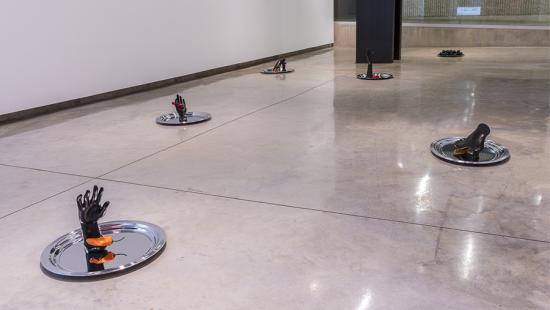 Six silver platters on a cement floor with black hands and feet on each one along with a red or orange colored pepper.