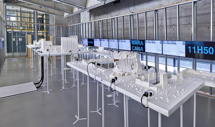 The modern interior of a gallery space, whose center is occupied by white tables displaying architectural models.