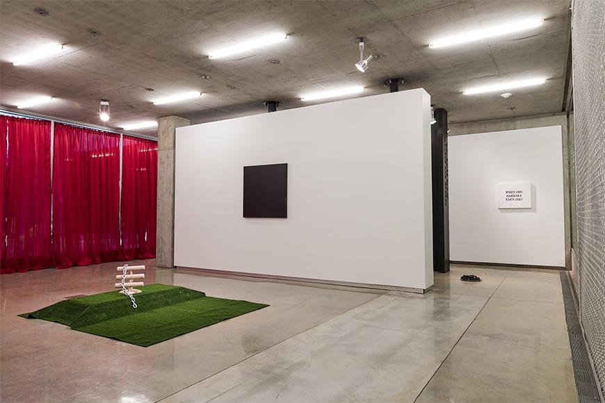 Red curtains in the back left, green turf with an abstract wooden sculpture on it, a white wall with a square dark image and another white wall with a small white sign with black letters.
