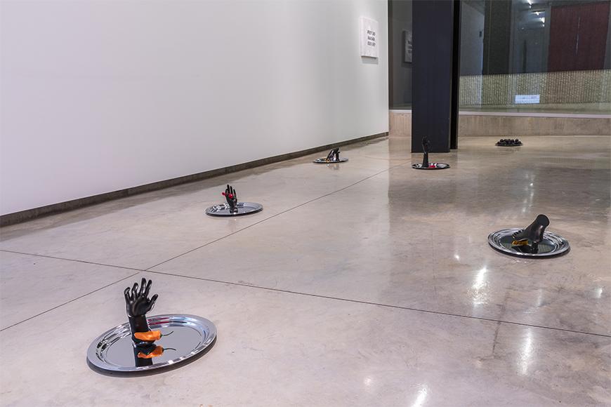 Six silver platters on a cement floor with black hands and feet on each one along with a red or orange colored pepper.