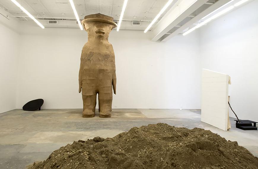 Large sculpture piece in a white room with dirt in front of it.