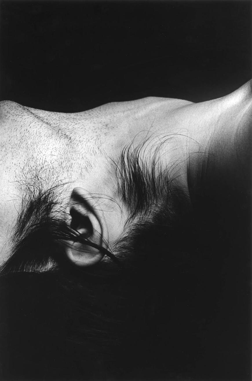 Black and white photograph of a person's neck and ear, with black hair tendrils surround the ear and black stubble on the cheek and chin.