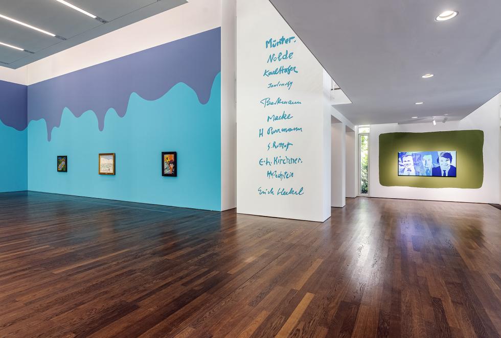 Large painted walls with smaller paintings hung next to a white wall with blue words on dark wooden floors.