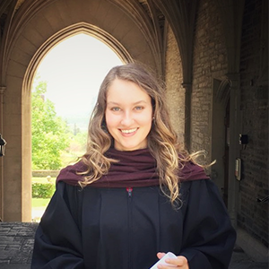 Portrait of Hailey Morey headshot of woman in black robe and brown scarf with arched entryway background