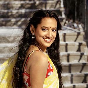 Girl with long dark hair smiling at the camera in a yellow and red shirt with stairs in the background.