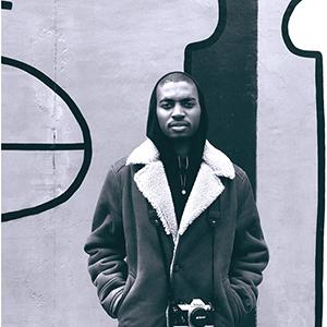 Black and white photo of a dark skinned person with masculine facial features wearing a black hoodie and grey winter coat standing in front of an abstract black and grey mural background
