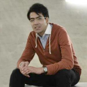 Portrait of Mitsuto Mori seated man with dark hair wearing a orange shirt and dark pants